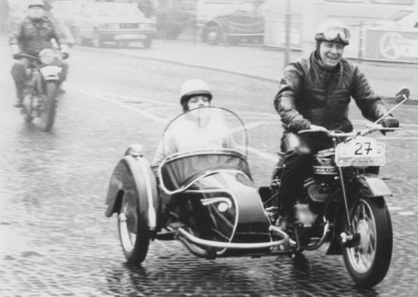 Zwei Männer fahren Motorrad einer sitzt auf der Maschine, der zweite im Beiwagen.