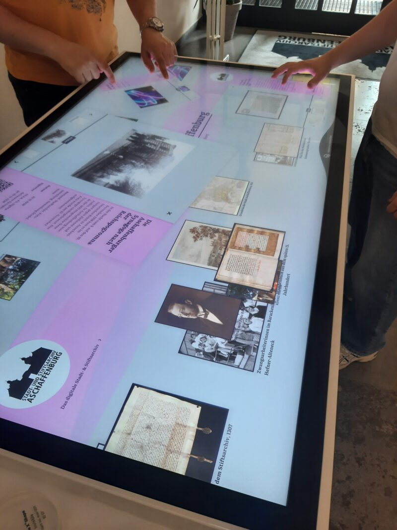 zwei Leute stehen am Medientisch im Foyer des Archis und berühren die Oberfläche, dabei öffnenen sich digitalisierte Dokumente